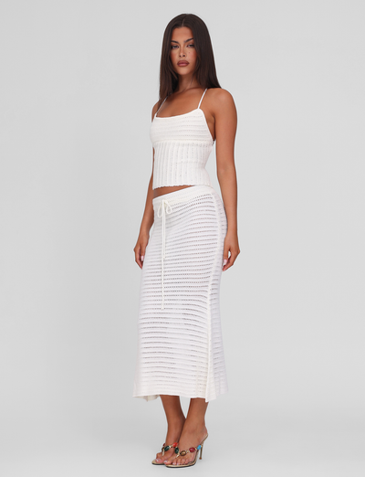 A woman with long brown hair wears the TIGERMIST EMELYN MIDI SKIRT in cream, featuring a crochet design and elasticated waist, paired with a white sleeveless ribbed knit top and heeled sandals against a plain background.