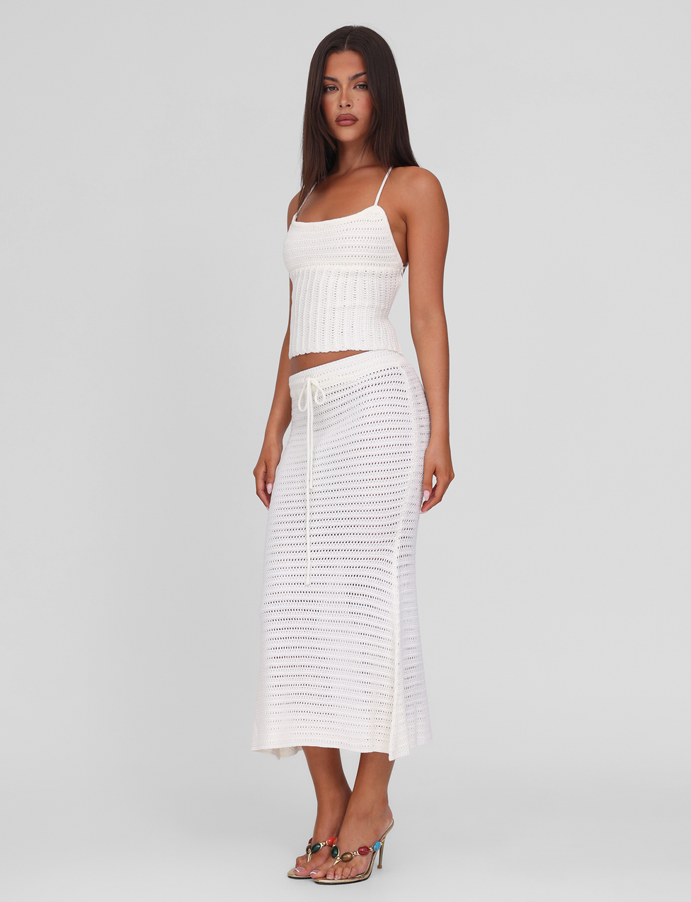 A woman with long brown hair wears the TIGERMIST EMELYN MIDI SKIRT in cream, featuring a crochet design and elasticated waist, paired with a white sleeveless ribbed knit top and heeled sandals against a plain background.
