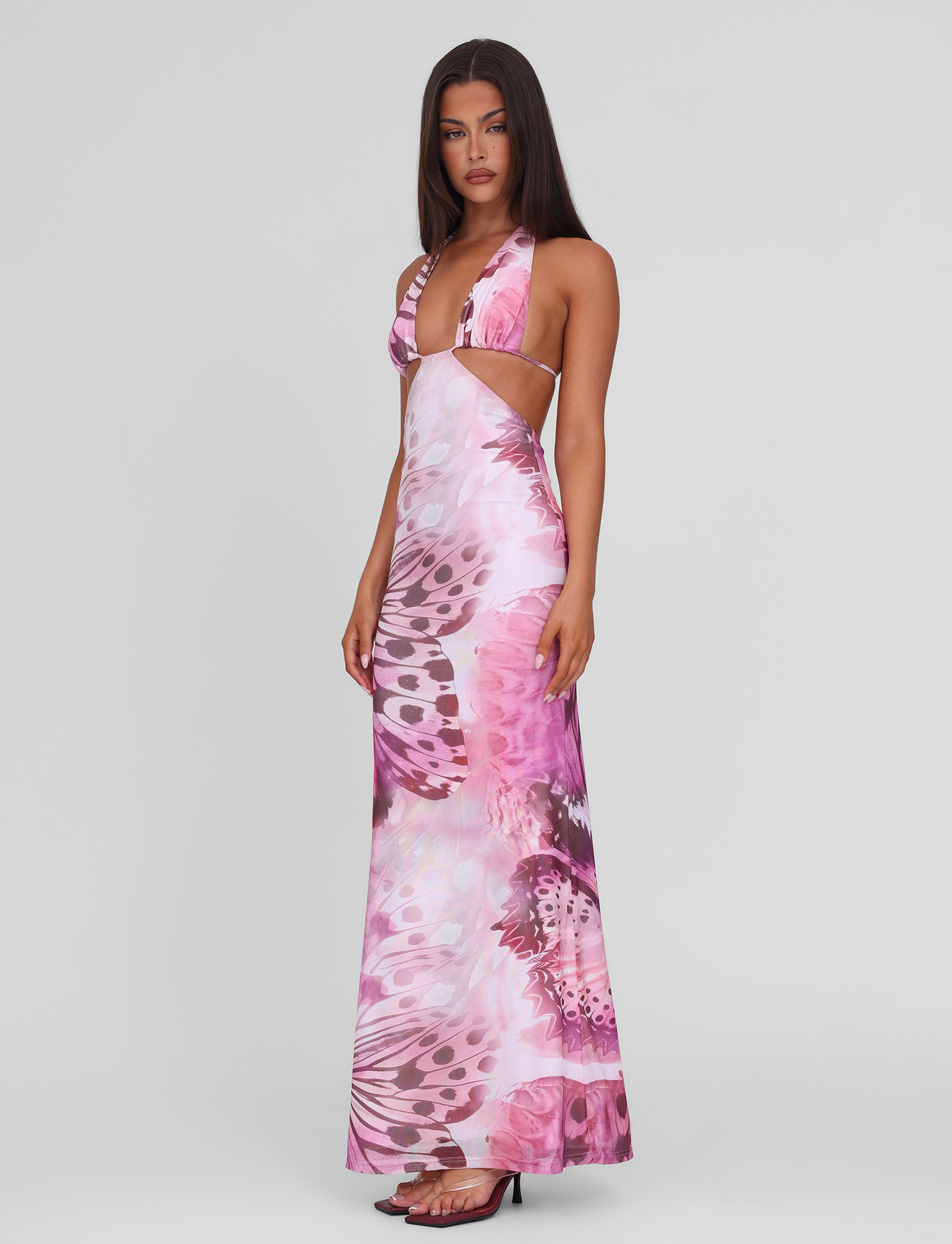 A woman poses against a plain background wearing the TIGERMIST SOLEIL MAXI DRESS - MACRO BUTTERFLY, featuring adjustable halter ties, side cutouts, and black heels.