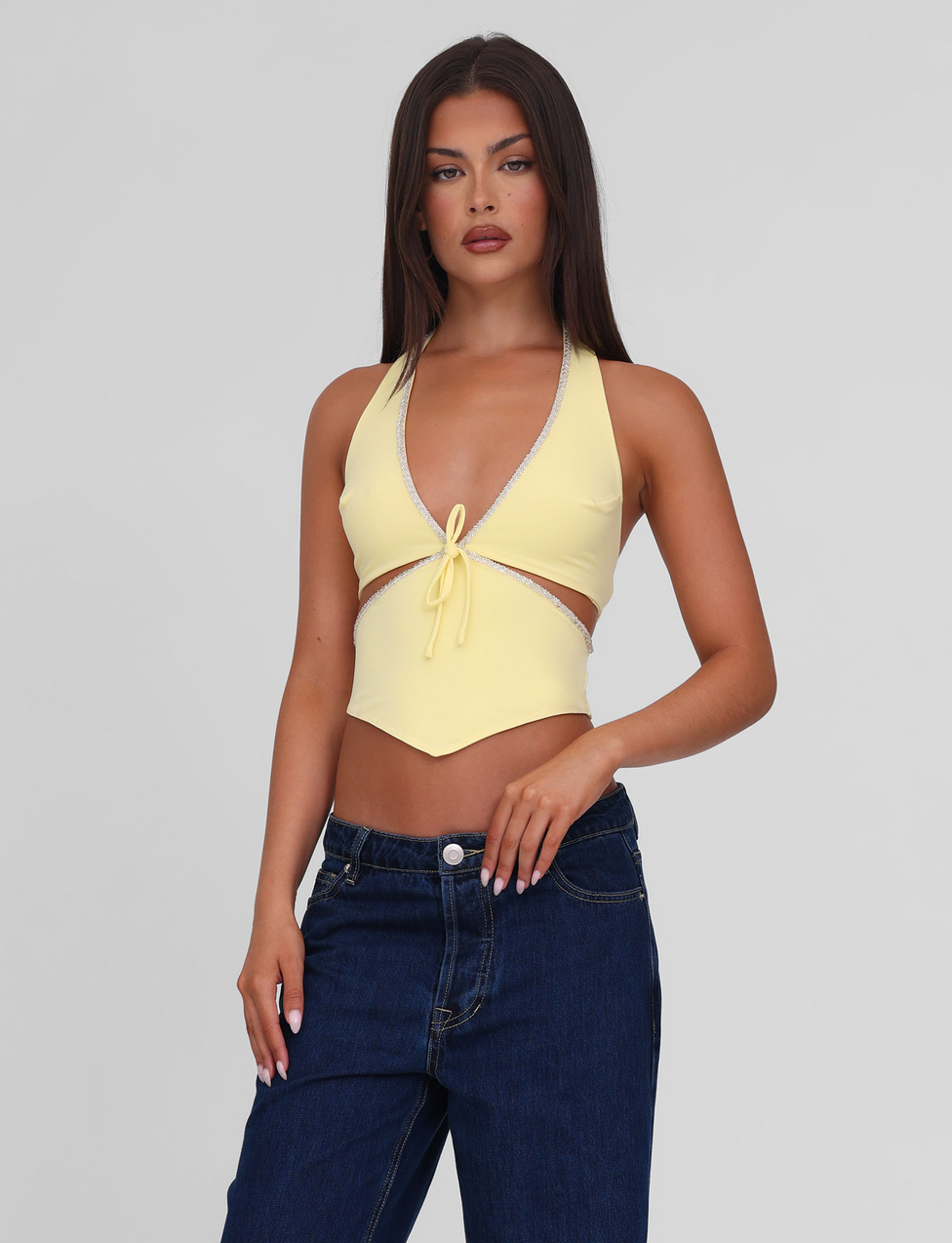 A woman with long dark hair models the TIGERMIST LIZZIE TOP - LEMON, a yellow halter style with a front tie and beaded trim, paired with blue jeans against a light gray background.