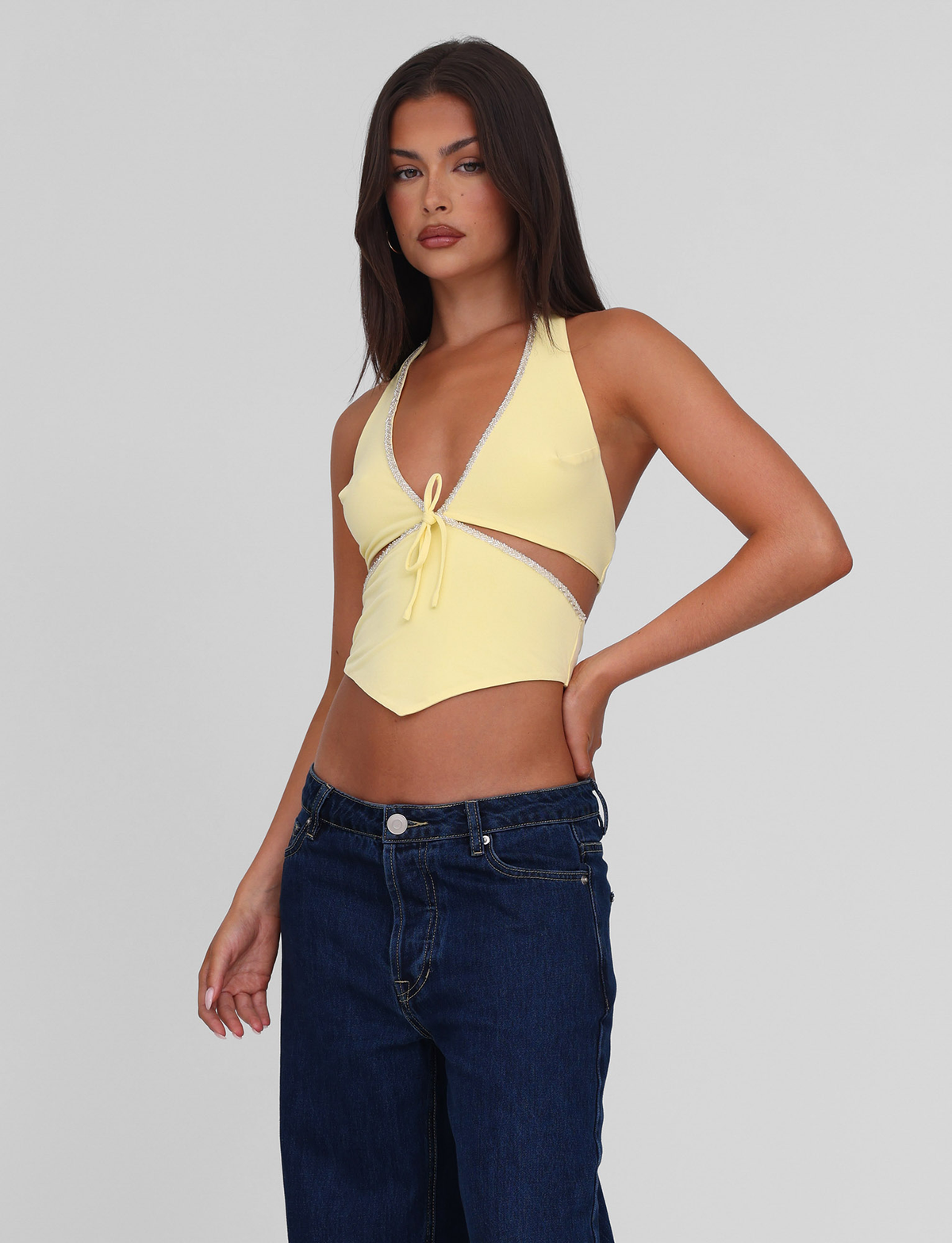 A woman with long brown hair models the TIGERMIST LIZZIE TOP - LEMON, a light yellow halter with cut-out details, paired with dark blue jeans. She stands against a plain light gray background, one hand on her hip.