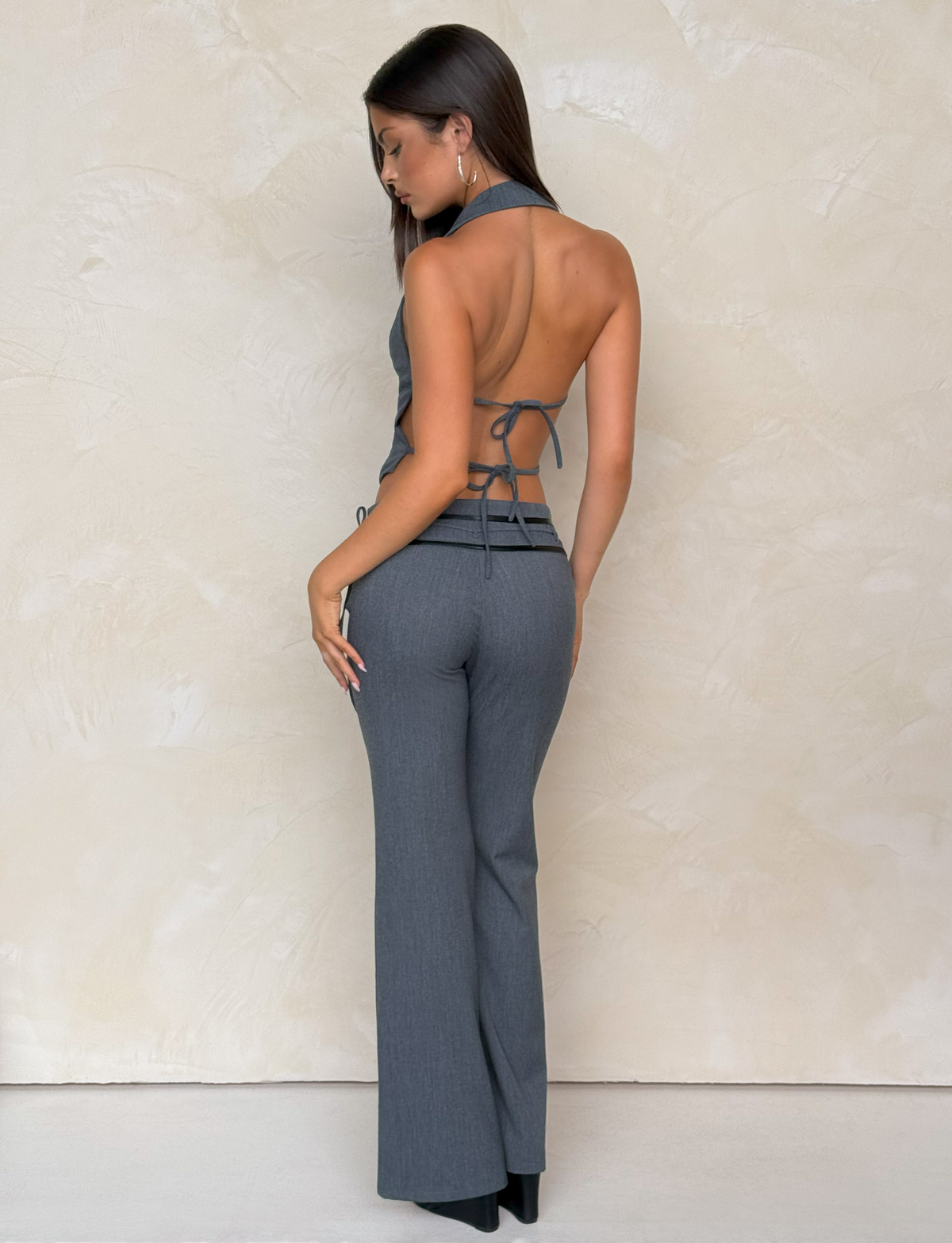 A woman with long straight brown hair wears TIGERMIST's FEDRA PANT - GREY, featuring a high-waisted tailored fit, paired with a backless halter top with tie closures, standing against a light beige textured background.