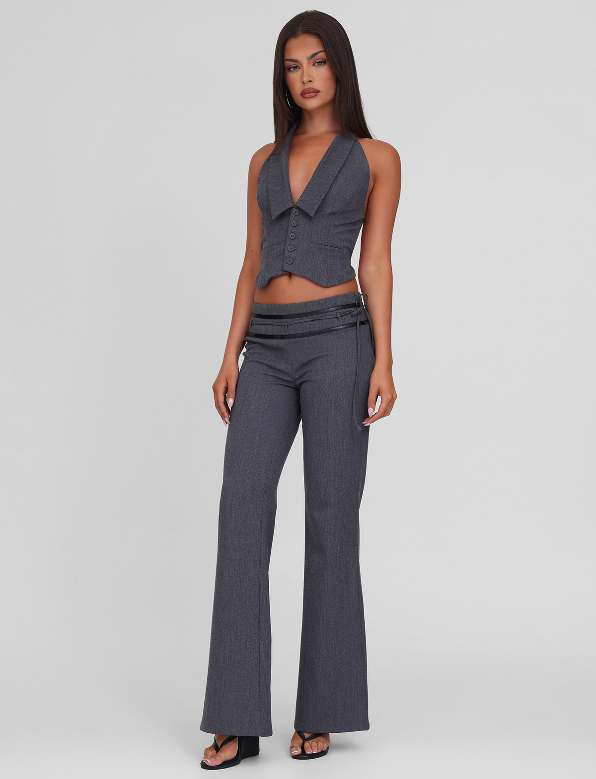 A woman with long dark hair wears the TIGERMIST FEDRA PANT - GREY, a pair of mid-rise tailored suit pants with side zippers, styled with a sleeveless deep V-neck gray vest and black high-heeled sandals against a light gray background.