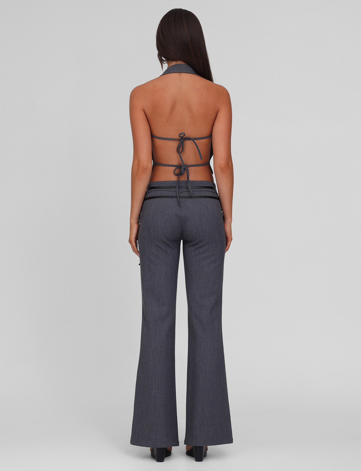 A woman with long dark hair faces away, wearing the TIGERMIST FEDRA PANT - GREY with a matching grey halter-style backless top tied with thin straps, set against a plain light grey background.