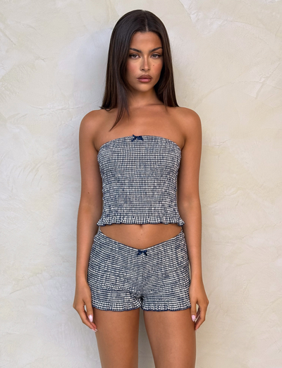 A woman with long straight brown hair stands before a light textured wall, wearing the TIGERMIST ERYNN TOP - NAVY GINGHAM and matching high-waisted bodycon shorts. She looks directly at the camera with a neutral expression.