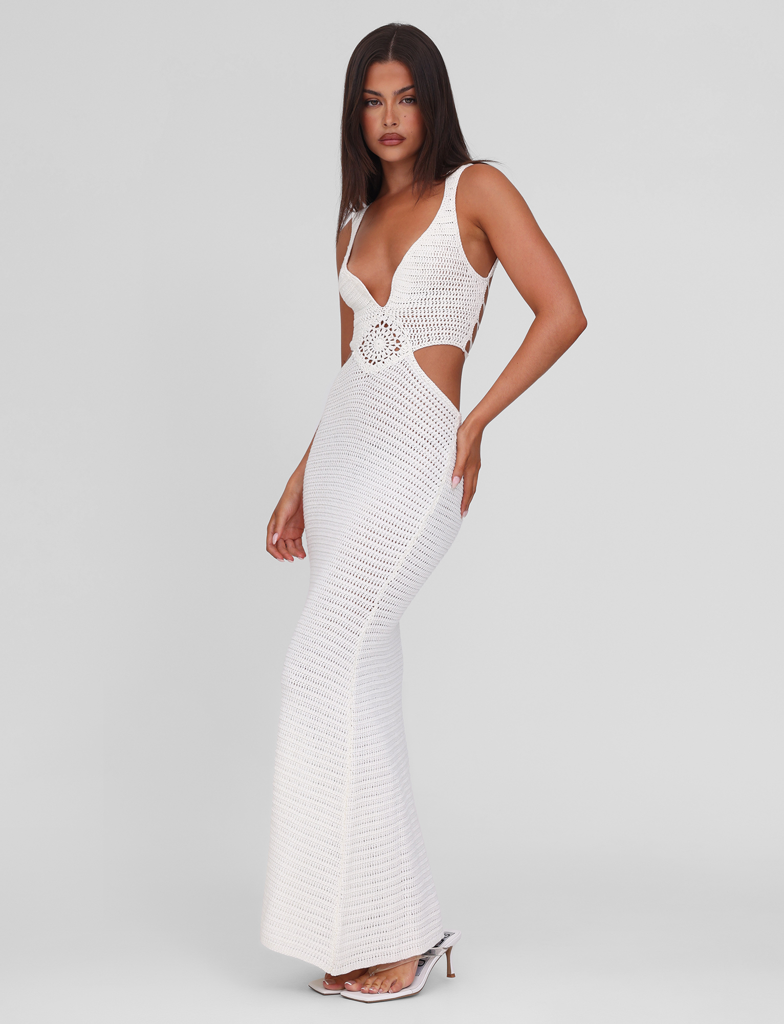 A woman in profile wears the TIGERMIST JONAH MAXI DRESS - CREAM, a bodycon, sleeveless crochet maxi with side cut-outs and deep neckline, styled with white open-toe heels against a plain light background.