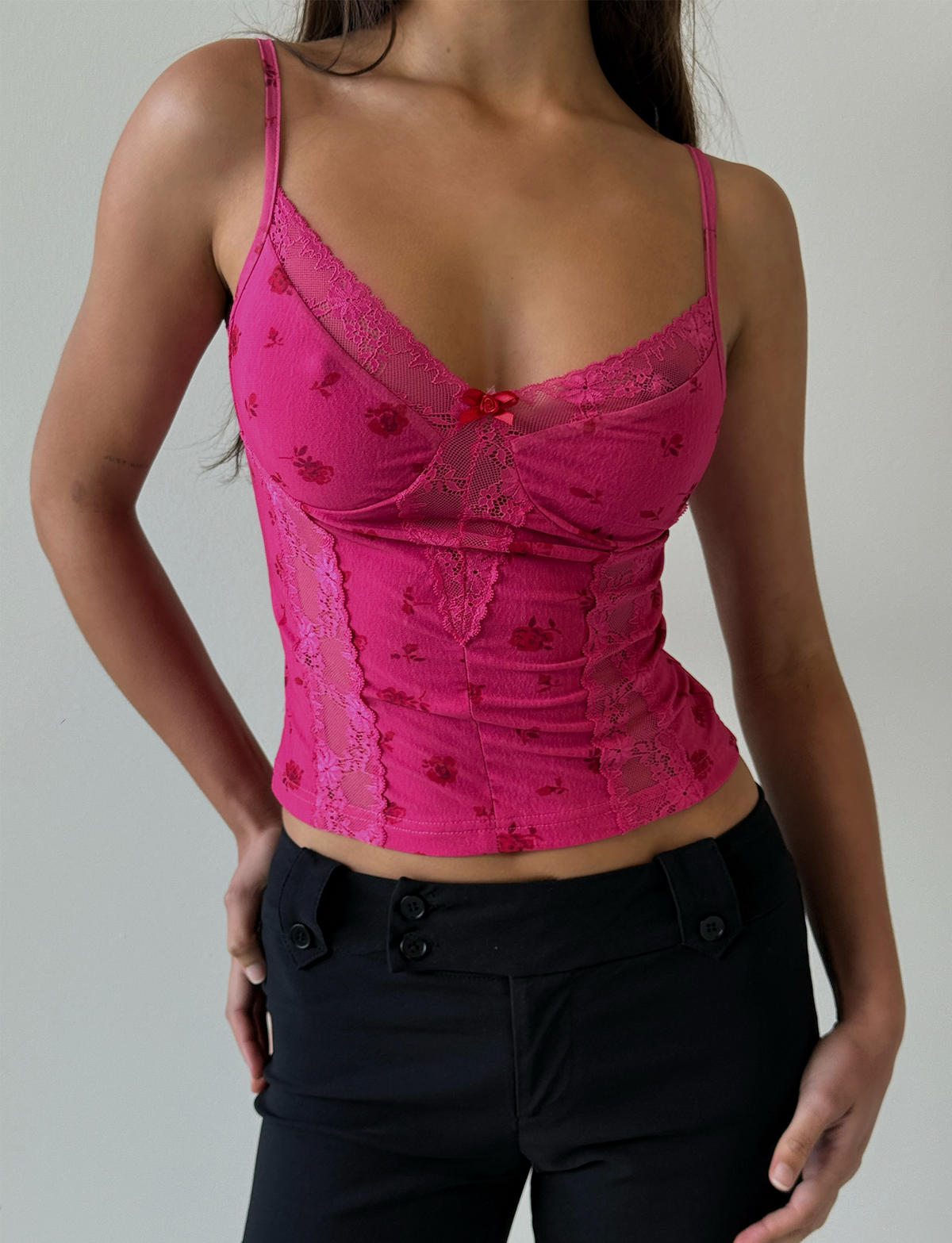 A woman wears the TIGERMIST KHALIA TOP - DITSY FLORAL, a fitted pink floral mesh camisole with lace trim, paired with black pants. She stands against a plain light background, hands on hips, and her head partially out of frame.
