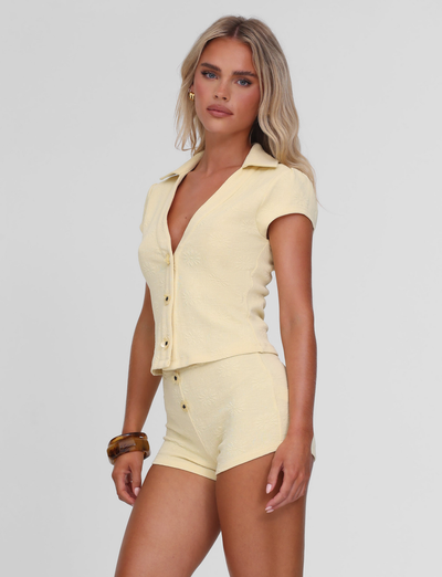 A woman with long blonde hair wears the TIGERMIST QUINLAN SHORT in lemon with a matching pale yellow button-up top, accessorized with a brown bangle. She stands against a plain white background, looking at the camera.