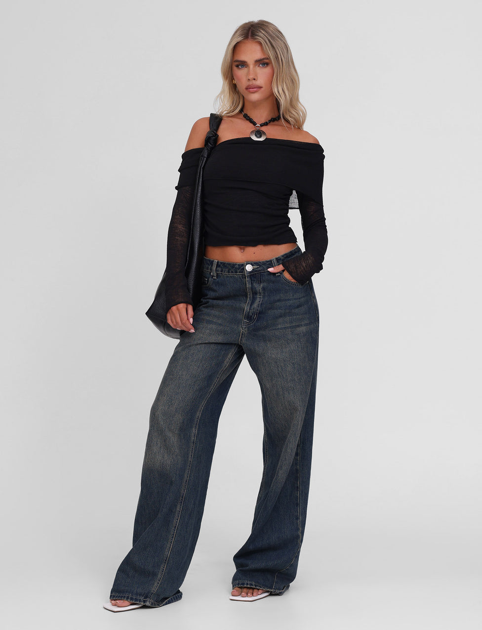 A woman with blonde hair poses confidently against a plain background, wearing the TIGERMIST VERA TOP in black, wide-leg blue jeans, clear-heeled sandals, and a black shoulder bag.
