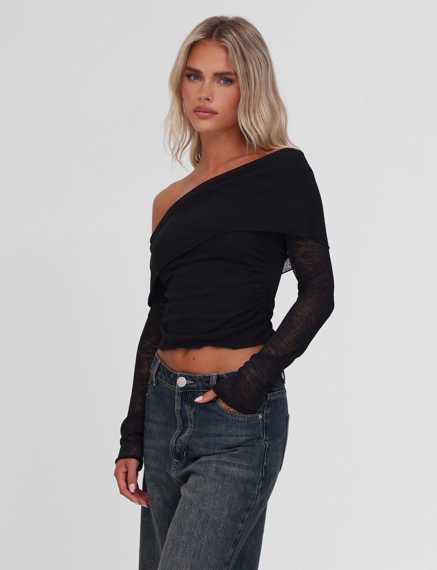 A person with long blonde hair models the TIGERMIST VERA TOP - BLACK, an off-shoulder knit with sheer long sleeves, paired with high-waisted blue jeans against a plain white background.
