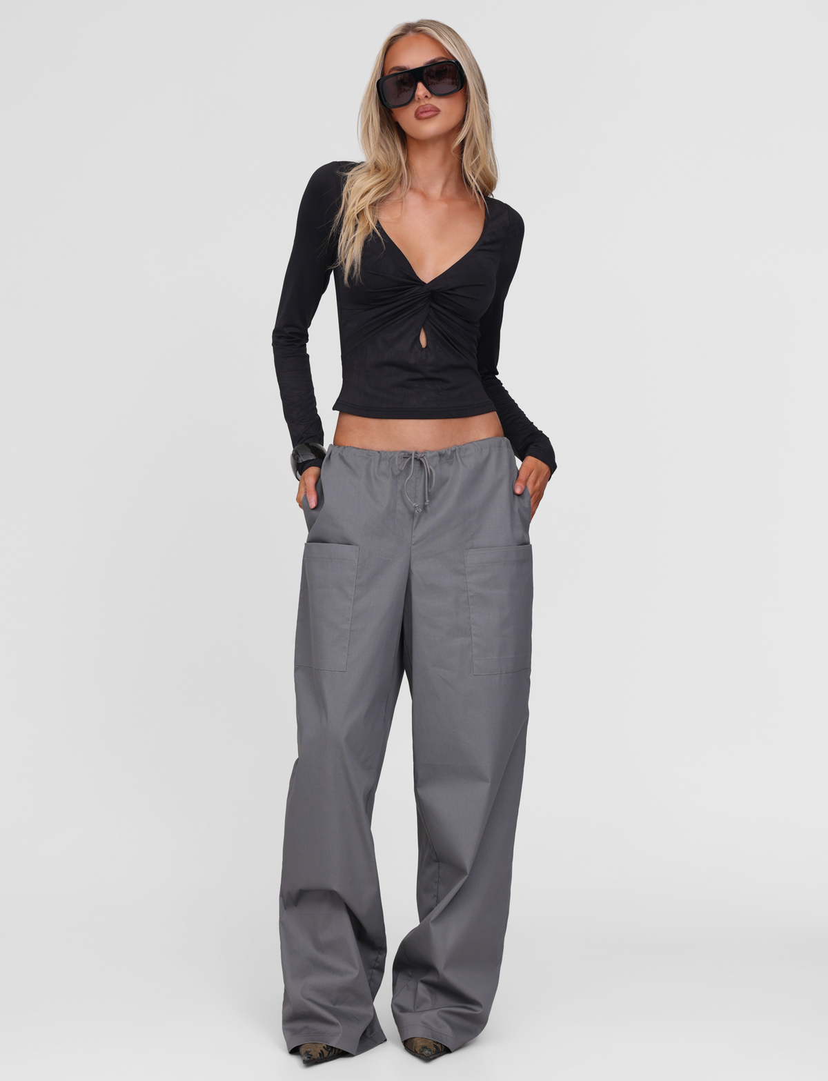 A woman in dark sunglasses, a black long-sleeve deep V-neck top, and TIGERMIST TYSHA PANT - CHARCOAL stands confidently with her hands in her pockets against a plain white background.