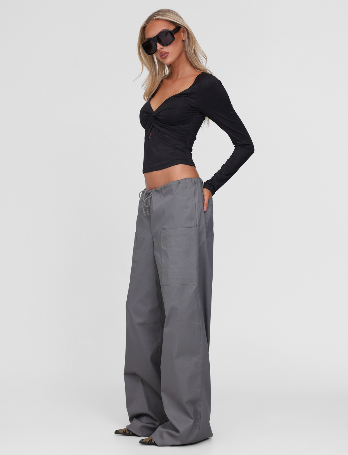 A woman stands slightly to the side against a plain white background, wearing large black sunglasses, a long-sleeve black crop top, and TIGERMIST TYSHA PANT - CHARCOAL wide leg cargo pants with a drawstring waist.