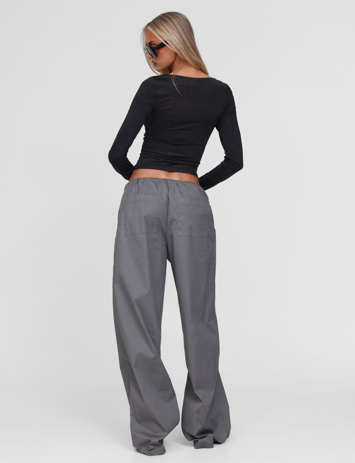 A woman with long blonde hair and sunglasses models the TIGERMIST TYSHA PANT in charcoal, featuring a wide-leg cargo style and drawstring waist, paired with a fitted black long-sleeve top against a white background.