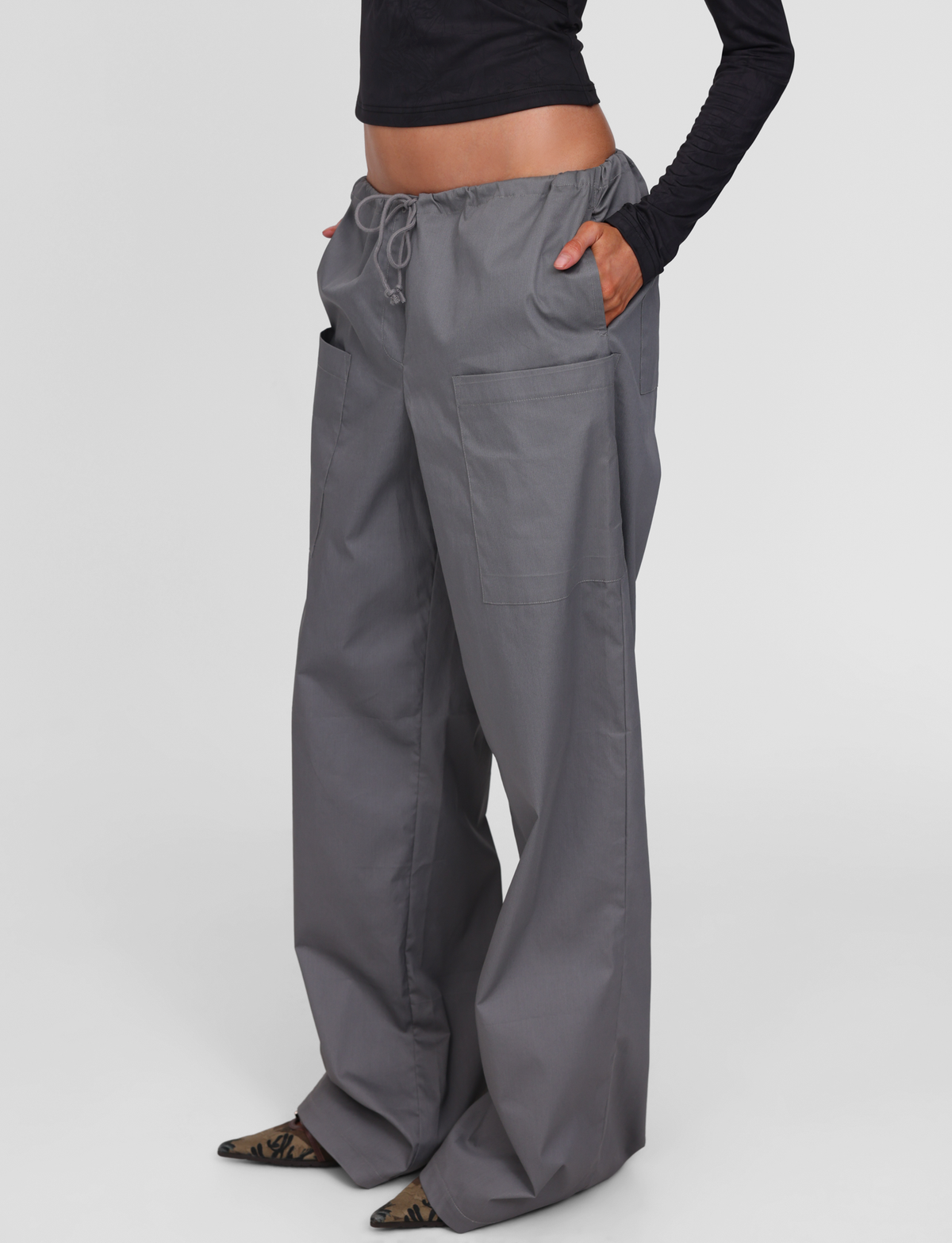 A person wears TIGERMIST’s TYSHA PANT in charcoal—wide leg cargo pants with a drawstring waist and large front pockets—styled with a cropped black long-sleeve top and pointed shoes, set against a plain white background.