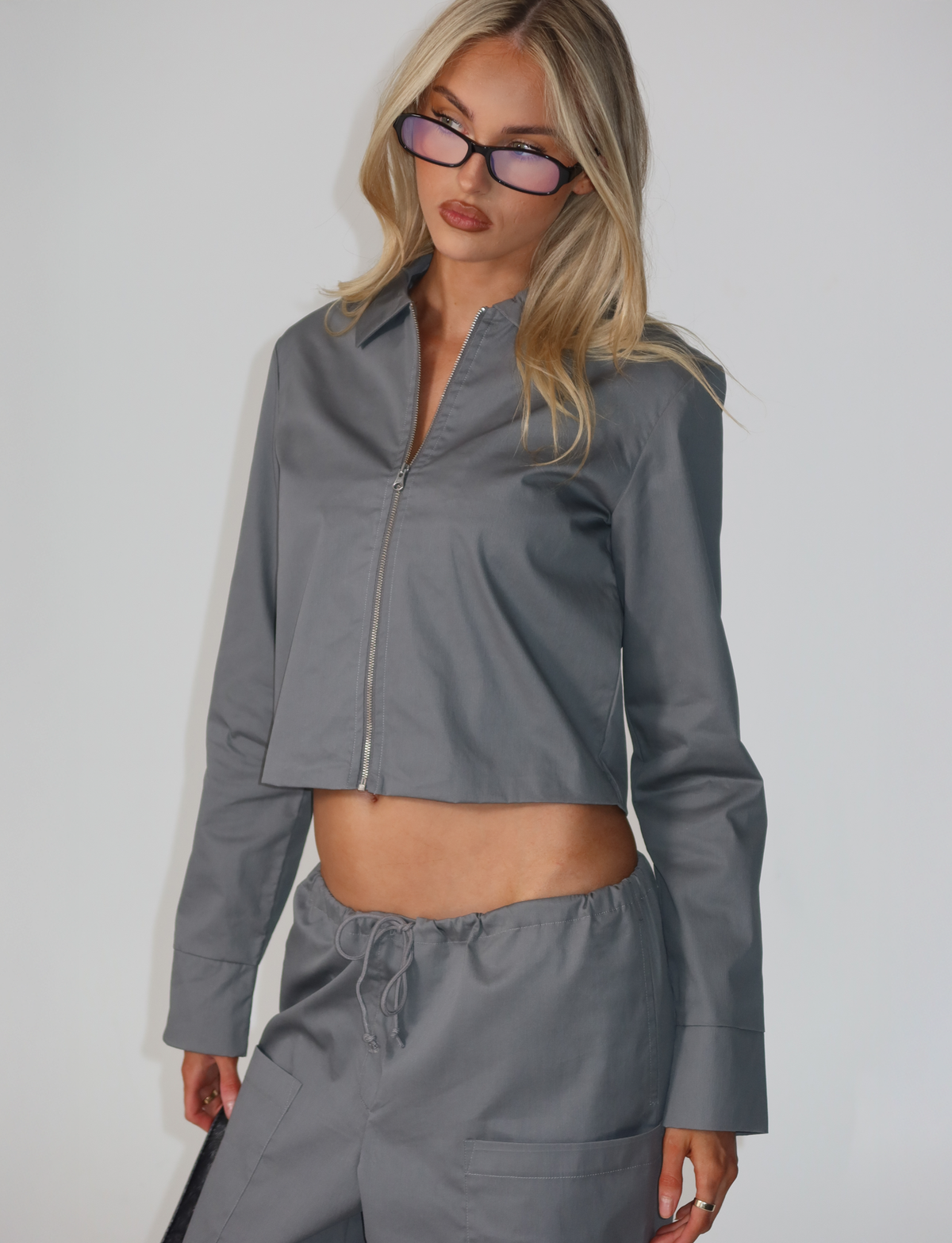 A woman with long blonde hair wears the TIGERMIST TYSHA JACKET in charcoal, styled with matching drawstring pants and sunglasses. She stands against a plain white background.