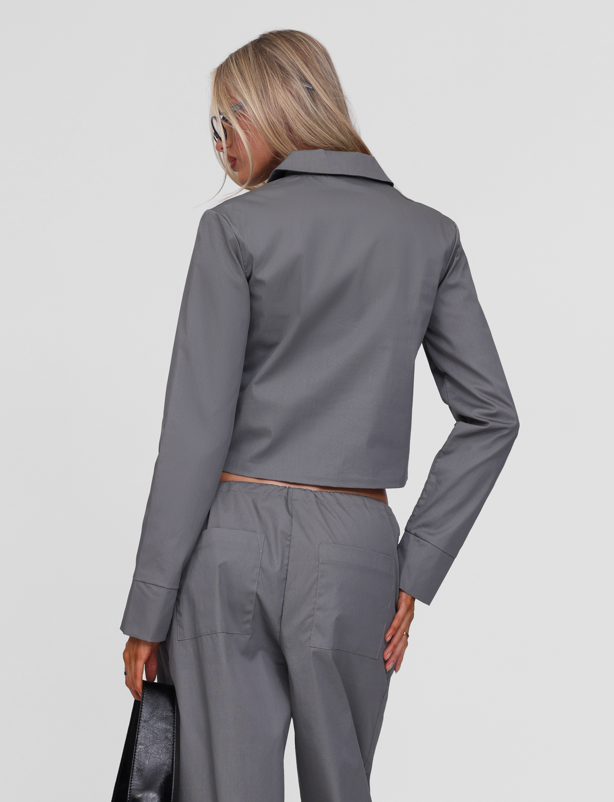 A woman with long blonde hair wears the TIGERMIST TYSHA JACKET in charcoal, paired with matching trousers, standing with her back to the camera while holding a black handbag and wearing sunglasses.
