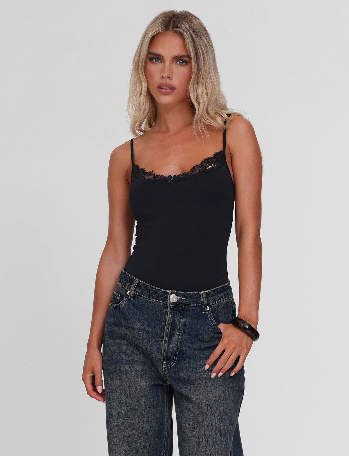 A woman with long blonde hair wears the TIGERMIST ASTRID BODYSUIT in black, paired with loose dark jeans, posing against a plain light background.