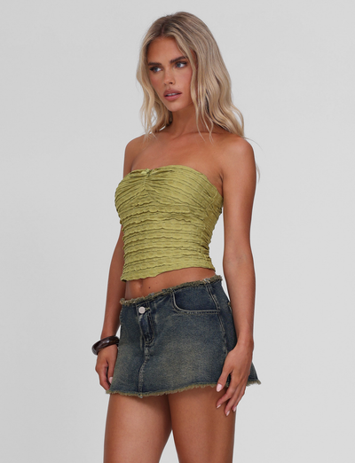 Woman with long blonde hair wears the TIGERMIST ROSALYN TOP - GREEN and a frayed dark denim mini skirt, standing against a plain white background.