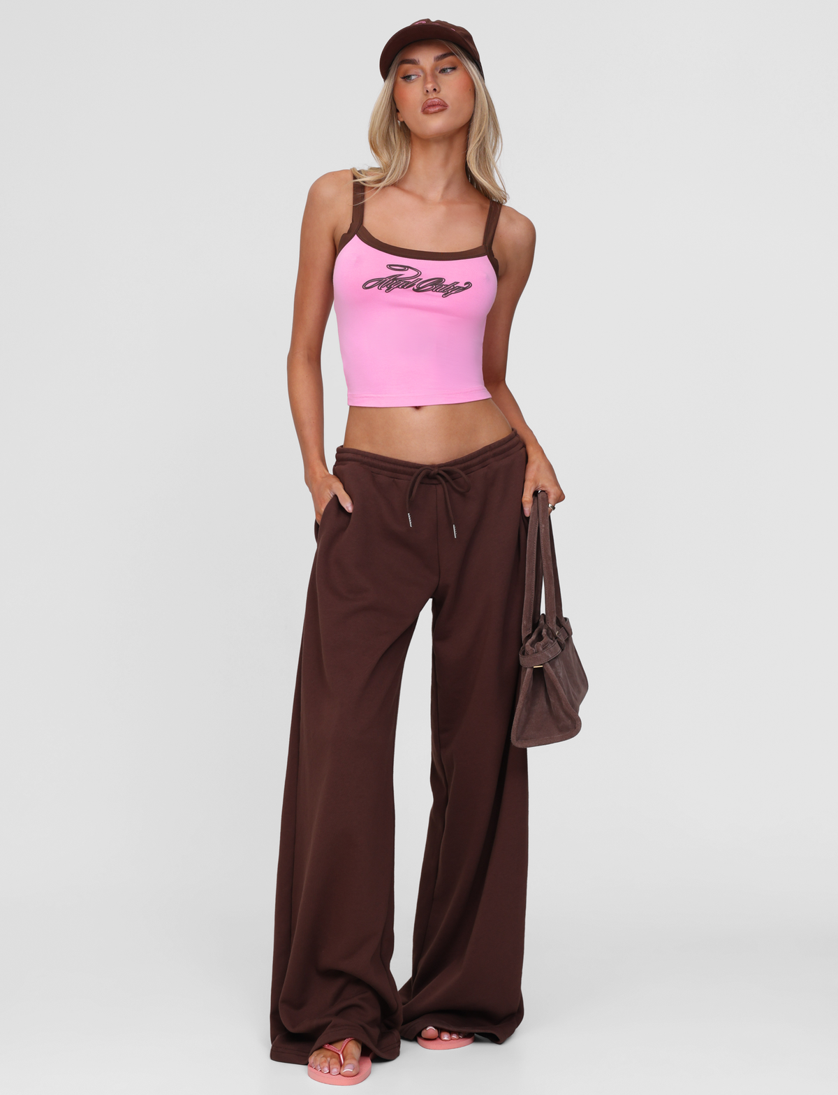 A woman wearing the TIGERMIST MERRYN TANK in pink with brown wide-leg pants, pink sandals, a brown cap, and a brown bag stands against a plain white background.
