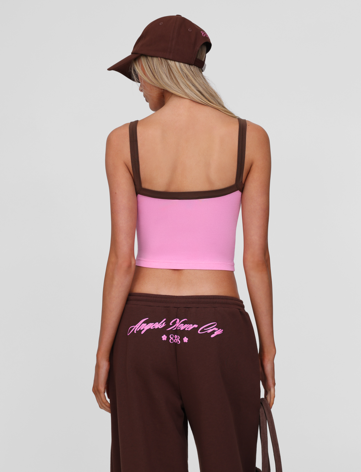 A person with long blonde hair, wearing a brown cap, brown pants with pink lettering on the back, and the TIGERMIST MERRYN TANK in pink is shown from behind against a plain light background.