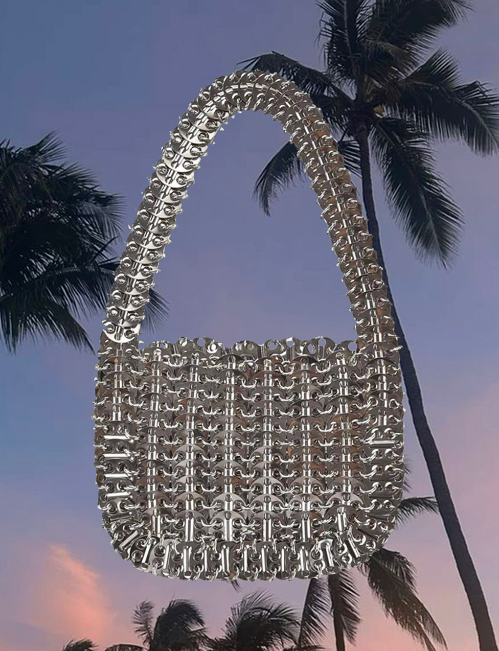 The TIGERMIST ZAZA BAG - SILVER, featuring a unique silver disc design and hand-held strap, is showcased against tall palm trees and a purple sunset sky.