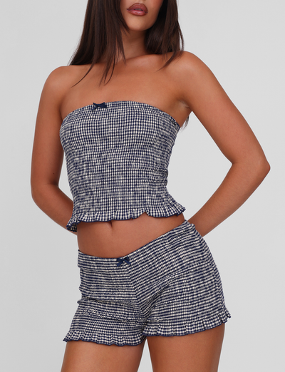 A woman wears the TIGERMIST ERYNN SHORTS in navy gingham with a matching strapless top, featuring small bow accents and a ruffled hem, set against a plain light background.