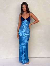 A woman with long dark hair stands against a light textured wall wearing the TIGERMIST ALAINA DRESS - BLUE ORCHID, featuring a blue floral print, V-neckline, and adjustable straps. She pairs it with white open-toe heels and a neutral expression.