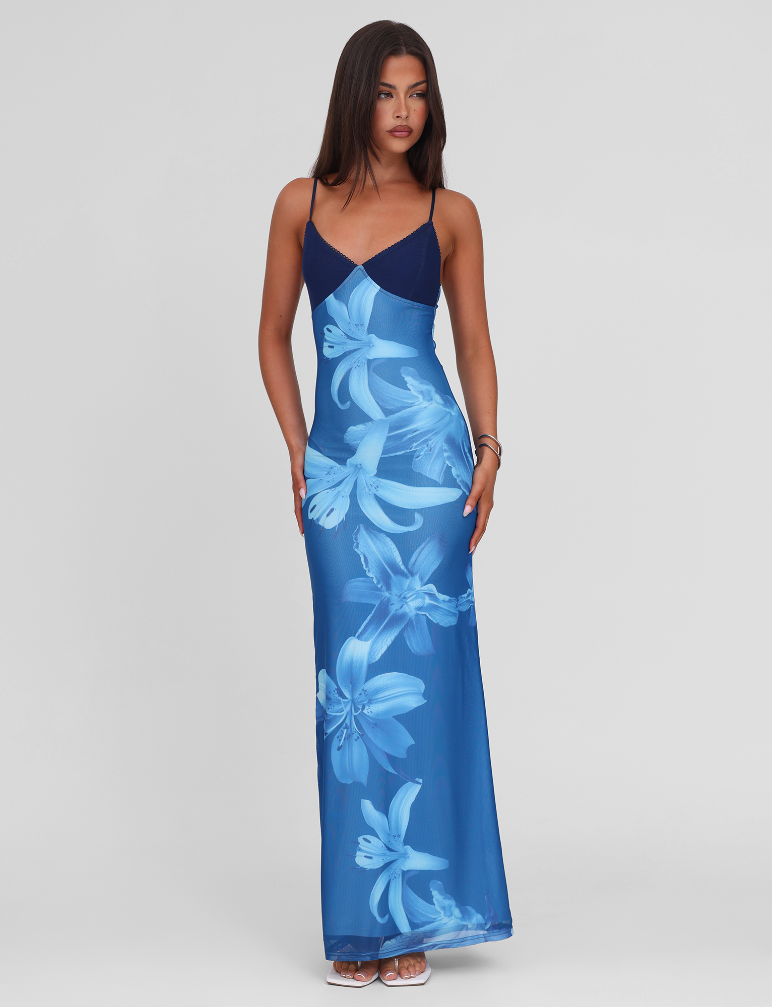 A woman with long dark hair wears the TIGERMIST ALAINA DRESS - BLUE ORCHID, a blue floral maxi dress featuring adjustable spaghetti straps and a V-neckline, styled simply with sandals against a plain background.