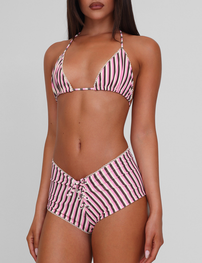 A woman wears the TIGERMIST DAZZLE BIKINI BOTTOM - STRIPE, featuring a pink, black, and white striped pattern with scallop lace detail and a lace-up high-waisted design, standing against a plain background.
