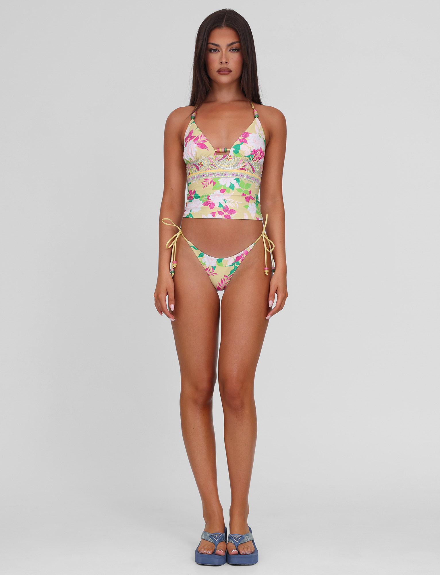 A woman with long brown hair and a neutral expression stands facing forward, wearing the TIGERMIST FINLEA TANKINI TOP - PAISLEY BORDER and matching bikini bottoms with yellow side ties, plus blue sandals, against a plain light gray background.