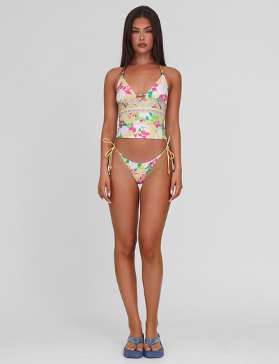 A woman with long brown hair and a neutral expression stands facing forward, wearing the TIGERMIST FINLEA TANKINI TOP - PAISLEY BORDER and matching bikini bottoms with yellow side ties, plus blue sandals, against a plain light gray background.