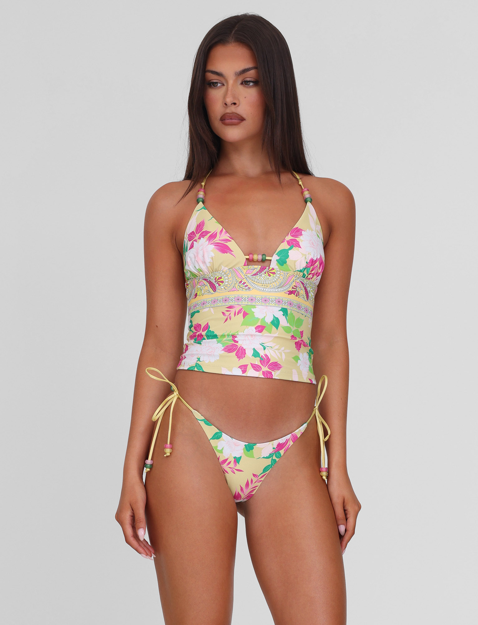 A woman with long brown hair models the TIGERMIST FINLEA BIKINI BOTTOM - PAISLEY BORDER, featuring tie-side cheeky bikini bottoms, paired with a tank-style top, standing against a plain light gray background.