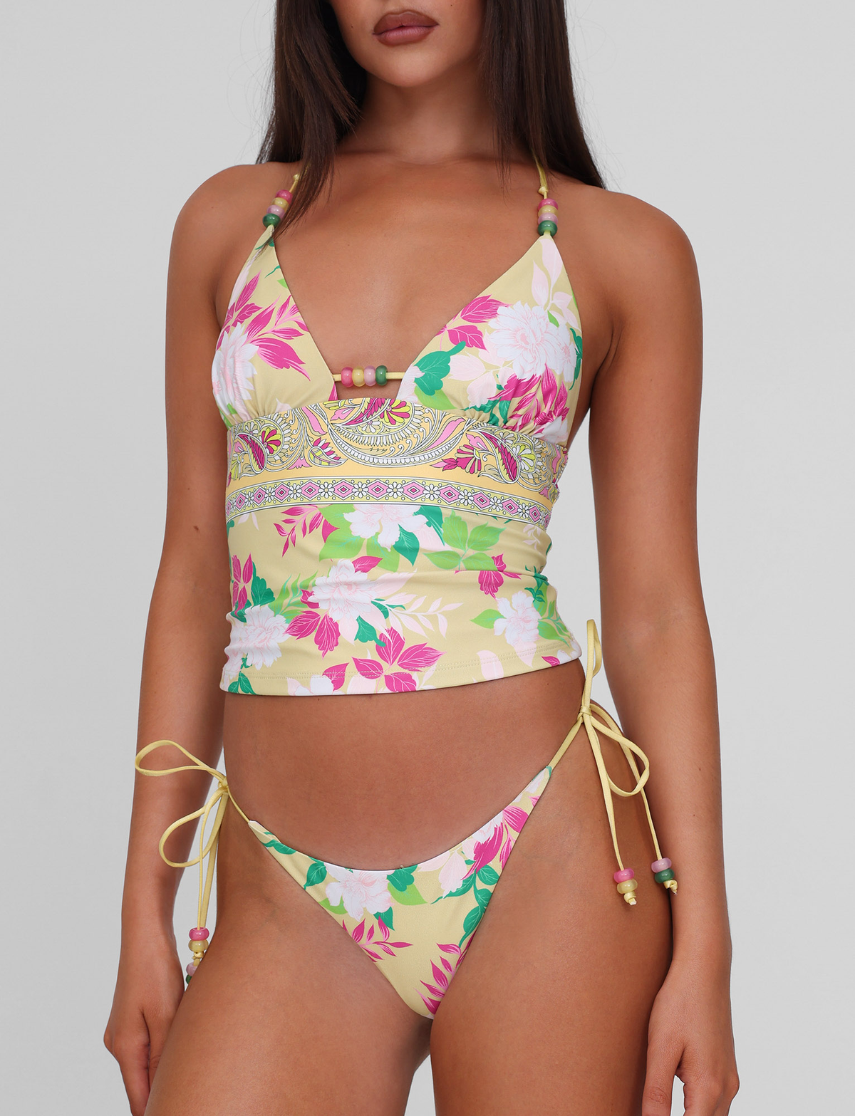 A woman wears the TIGERMIST FINLEA BIKINI BOTTOM - PAISLEY BORDER with a matching yellow floral tankini top, both featuring a cheeky bikini cut, standing against a plain light background.