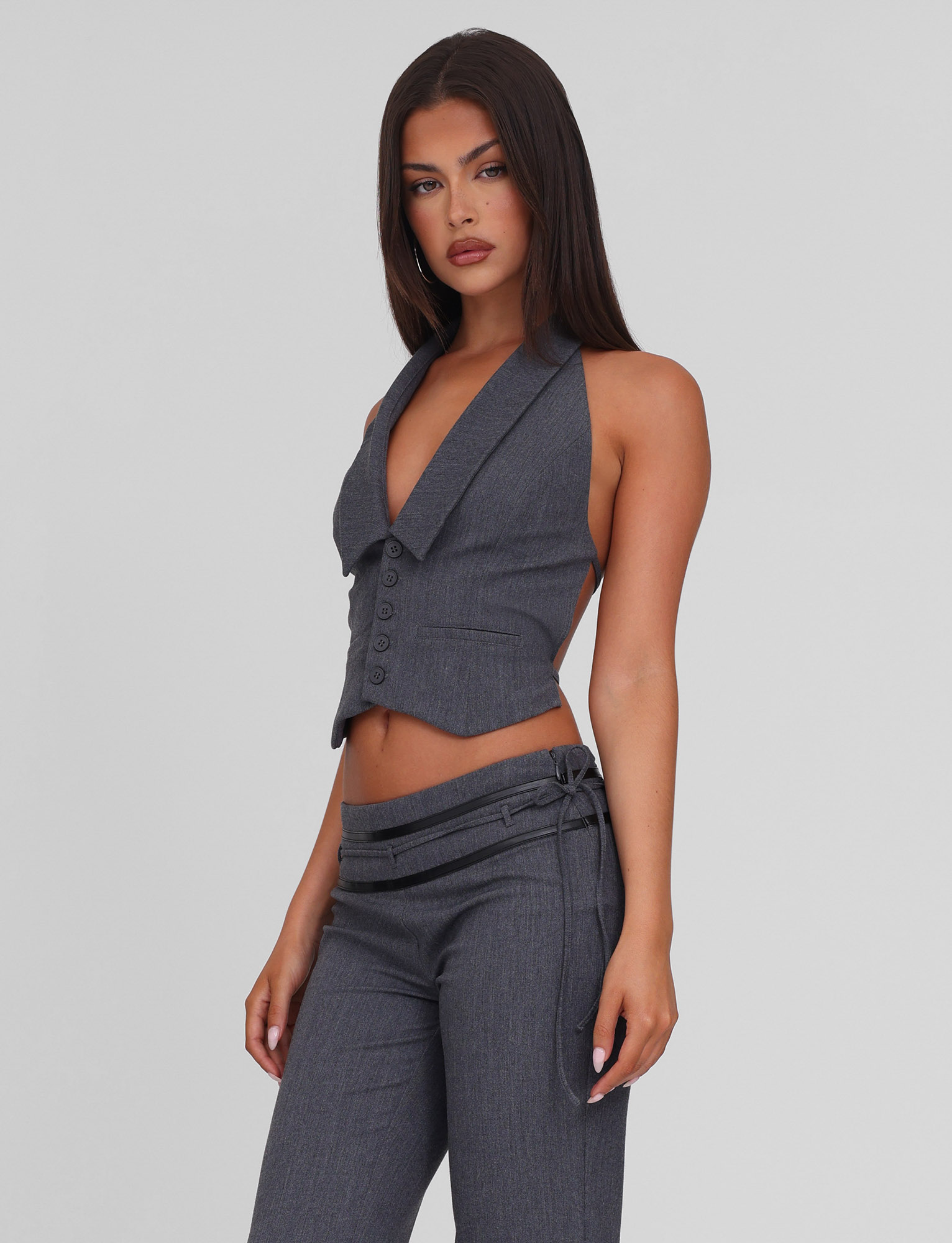 A woman with long straight hair and a neutral expression models the TIGERMIST FEDRA VEST - GREY, a tailored fit, deep V-neck vest with buttons, paired with matching low-waist gray pants featuring a decorative side tie.