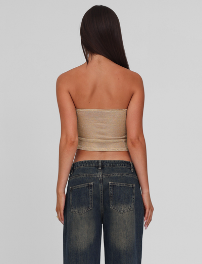 A woman with long brown hair faces away, wearing the TIGERMIST HARRIETT TUBE TOP in gold and wide-leg dark blue sparkle jeans, set against a plain light gray background.