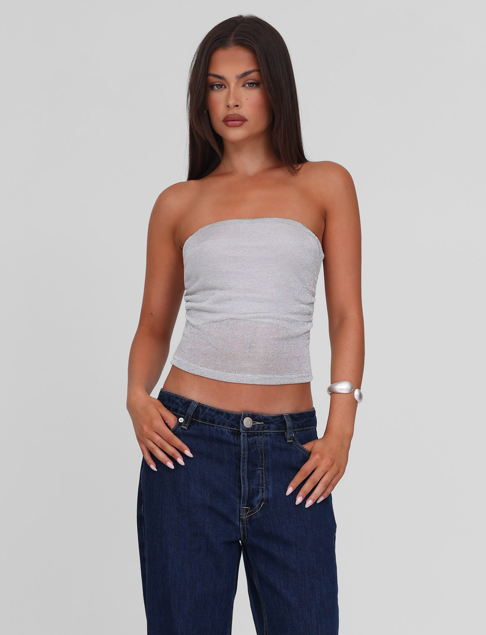 A woman with long dark hair wears the TIGERMIST HARRIETT TUBE TOP - SILVER, paired with dark blue jeans and a white bracelet. She stands against a plain light gray background with a neutral expression.