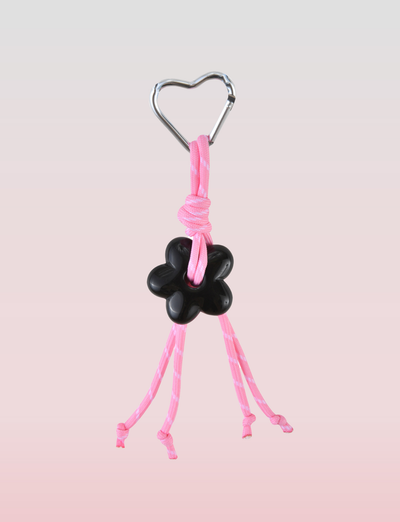 The TIGERMIST MIRIAM KEYCHAIN - HOT PINK features a metal heart clasp, pink cords with a black flower-shaped bead, and four loose ends, all set against a soft pink gradient—perfect as a stylish accessory.