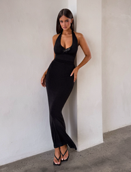 A woman with long dark hair wears the TIGERMIST ASTORIA MAXI DRESS in black and black sandals, standing indoors against a white wall on a polished concrete floor.