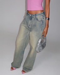 A person in a bright pink sleeveless top, pink open-toe heels, and the TIGERMIST ZANTHE JEAN - GREY WASH holds a metallic silver purse and wears a silver cuff bracelet against a plain light background.