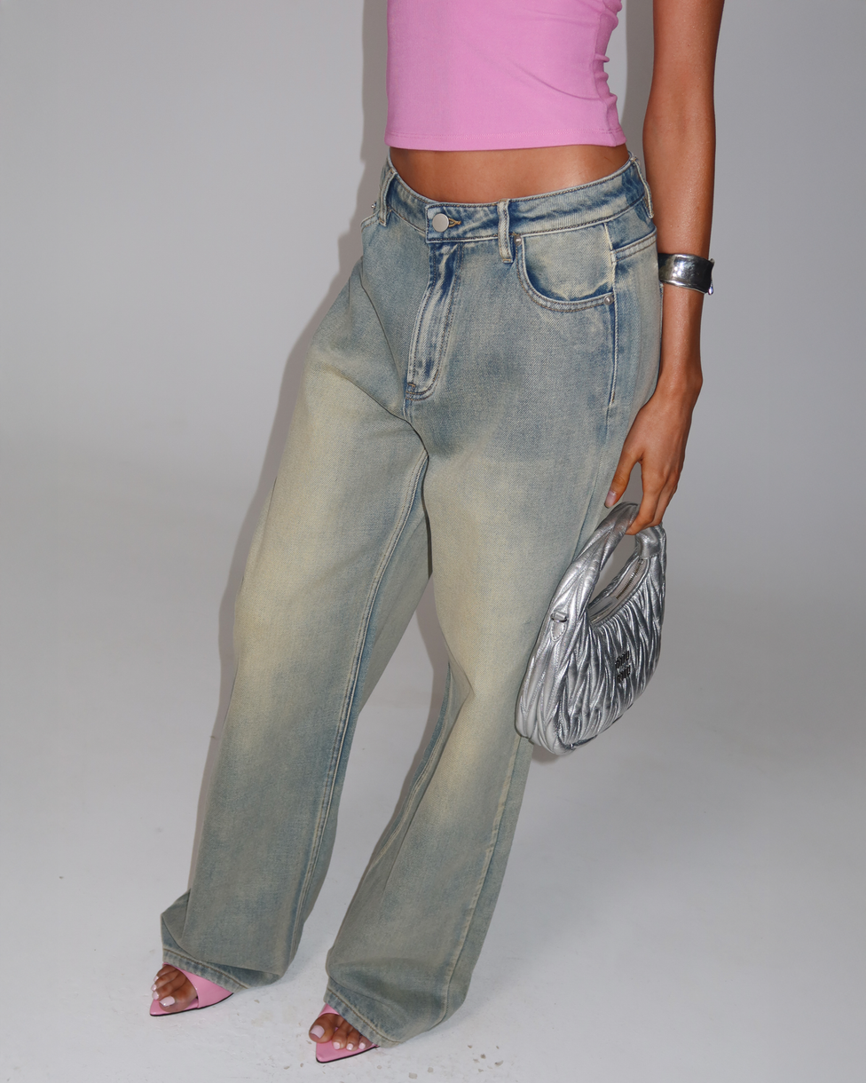 A person in a bright pink sleeveless top, pink open-toe heels, and the TIGERMIST ZANTHE JEAN - GREY WASH holds a metallic silver purse and wears a silver cuff bracelet against a plain light background.