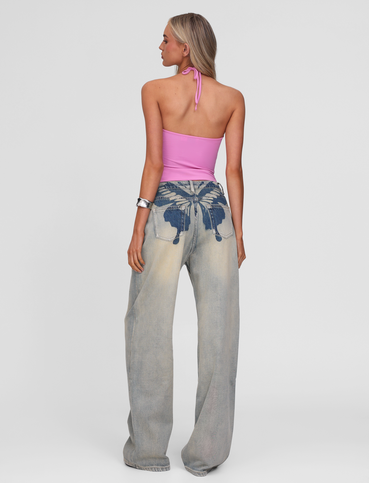 A woman with long blonde hair and silver bracelets wears the TIGERMIST ZANTHE JEAN - GREY WASH, featuring high-waisted, wide-leg, oversized fit with bold blue wing and heart graphics on the back pockets, paired with a pink halter top.