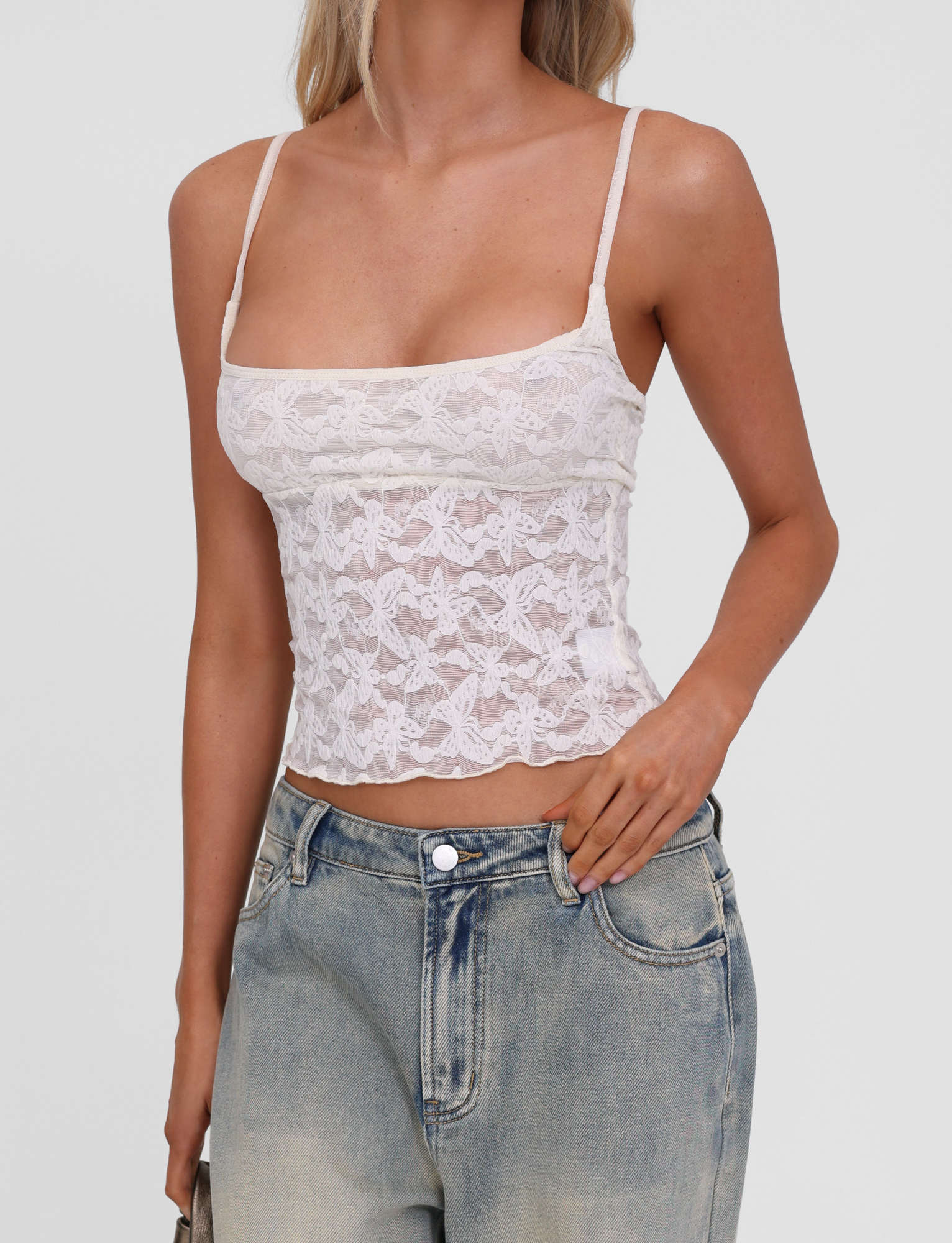 A woman wears the TIGERMIST GIULIA 2PC TOP in ivory lace, featuring adjustable straps and a butterfly pattern, paired with light blue high-waisted jeans. She poses with one hand in her pocket against a plain background.