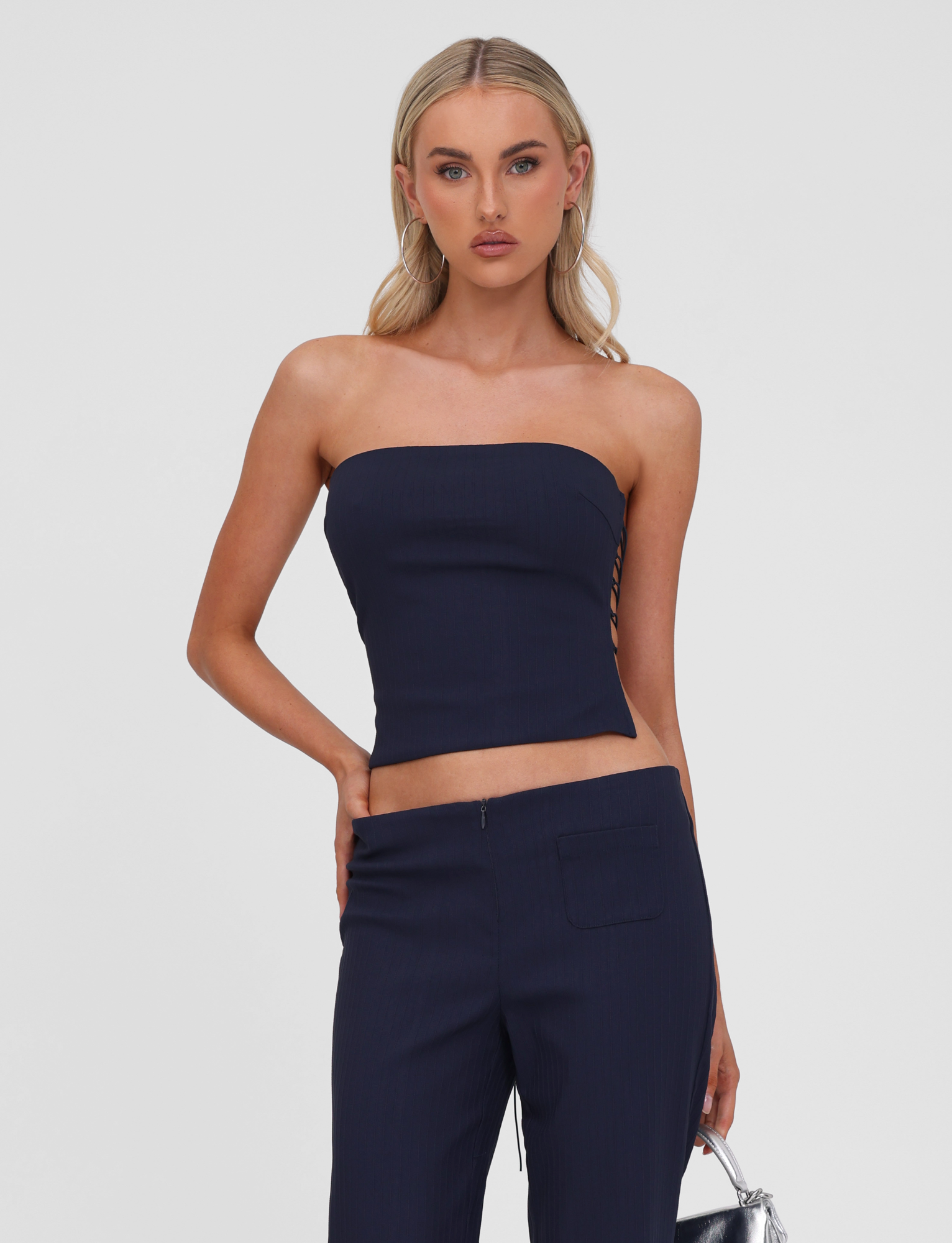 A woman with long blonde hair poses against a white background, wearing the TIGERMIST GWENDOLINE TOP in navy—a strapless style with a lace-up back—paired with matching pants, one hand resting on her hip.