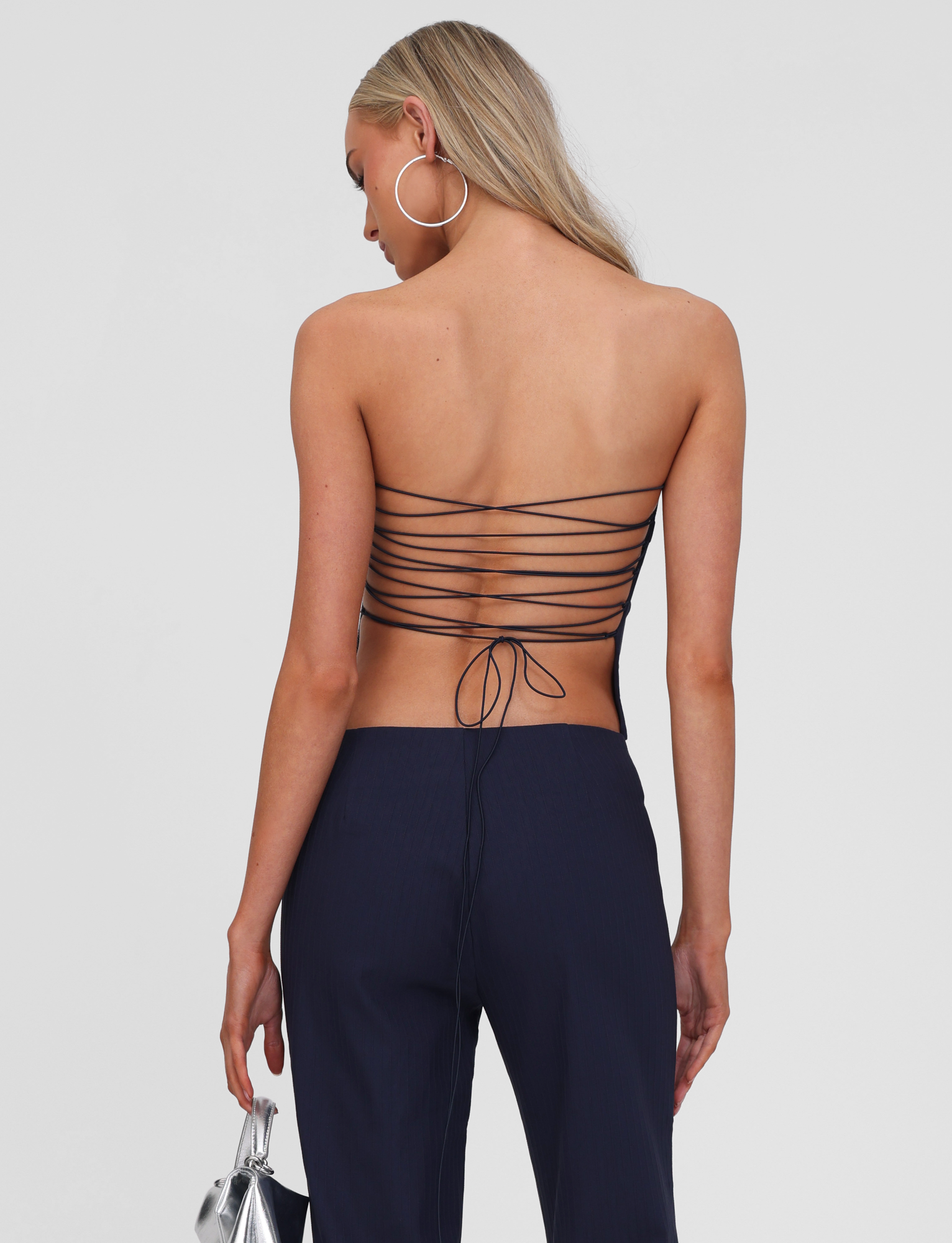 A woman with long blonde hair wears the TIGERMIST GWENDOLINE TOP - NAVY, featuring a lace-up back with thin crisscrossing straps, navy pants, large hoop earrings, and holds a small silver handbag. She is photographed from behind against a plain background.