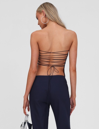 A woman with long blonde hair wears the TIGERMIST GWENDOLINE TOP - NAVY, featuring a lace-up back with thin crisscrossing straps, navy pants, large hoop earrings, and holds a small silver handbag. She is photographed from behind against a plain background.
