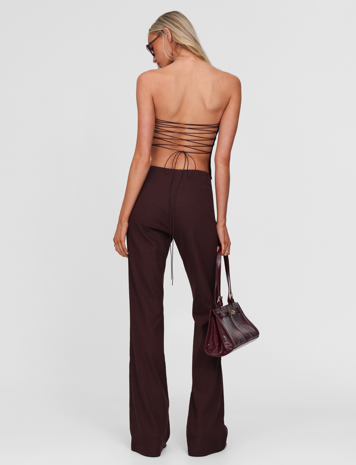 A woman with long blonde hair faces away, wearing TIGERMIST’s GWENDOLINE PANT - CHOCOLATE, a high-waisted flared suit pant. She pairs it with a backless strappy top, holds a dark brown handbag, and wears sunglasses against a plain white background.