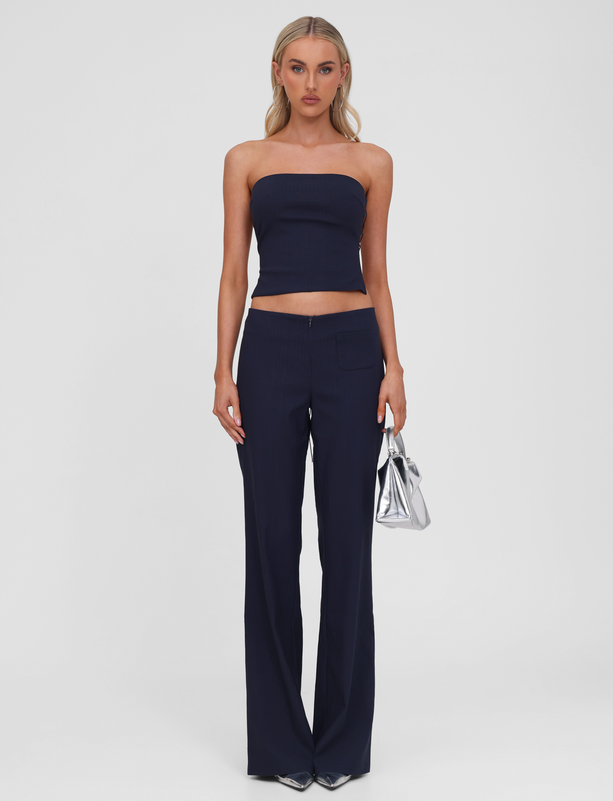 A woman poses against a white background, wearing the TIGERMIST GWENDOLINE PANT in navy with a matching strapless crop top. She accessorizes with a shiny silver handbag and metallic pointed-toe shoes.