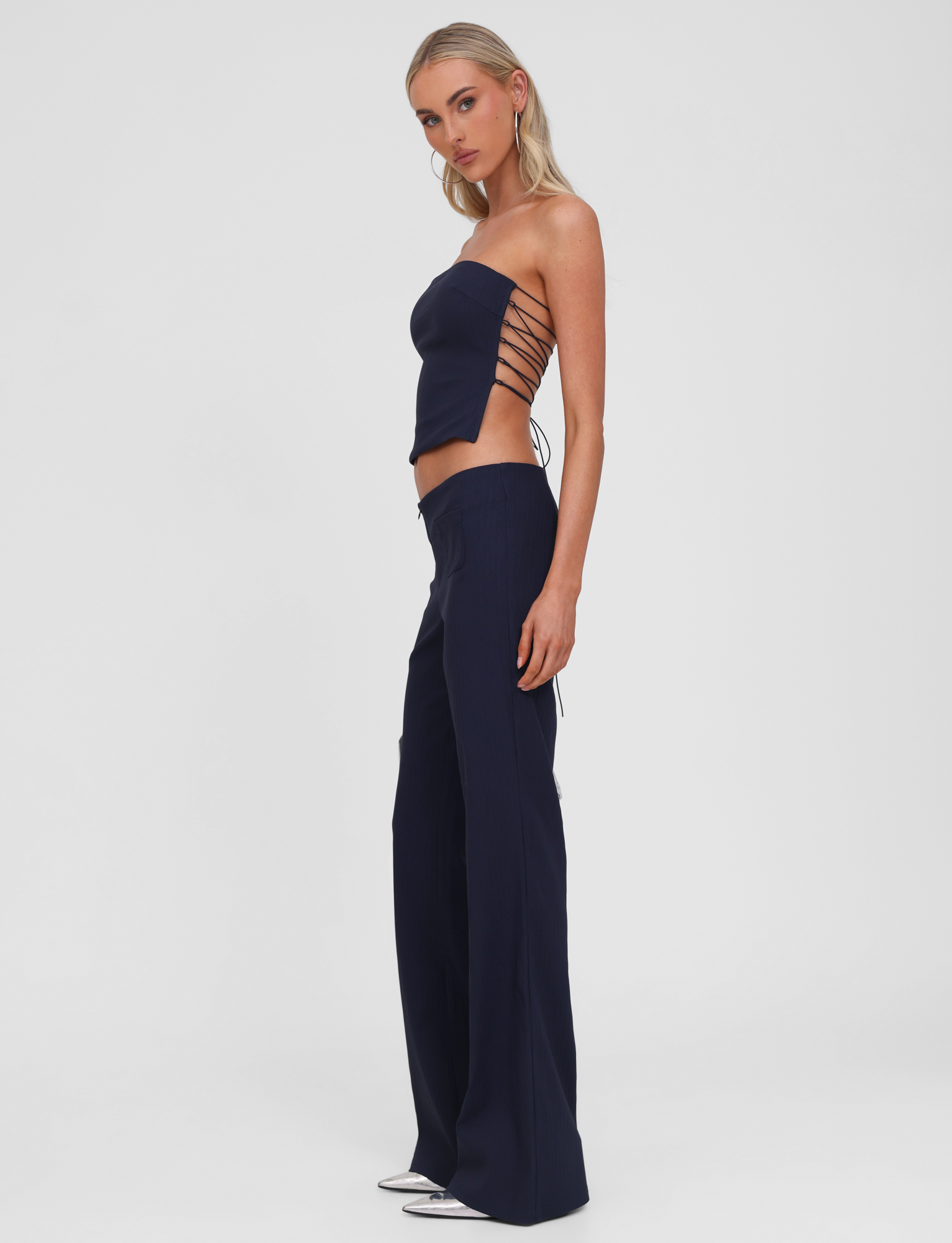 A woman models the TIGERMIST GWENDOLINE PANT in navy—low rise, flared leg suit pants—with a navy strapless top featuring crisscross side ties. She stands in profile against a white background, wearing silver pointed-toe shoes.