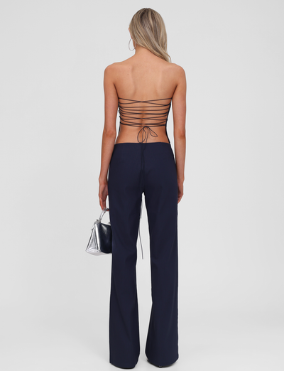 A woman with long blonde hair faces away, wearing a strappy backless top, TIGERMIST's GWENDOLINE PANT in navy flared leg style, and holding a metallic silver handbag against a plain white background.