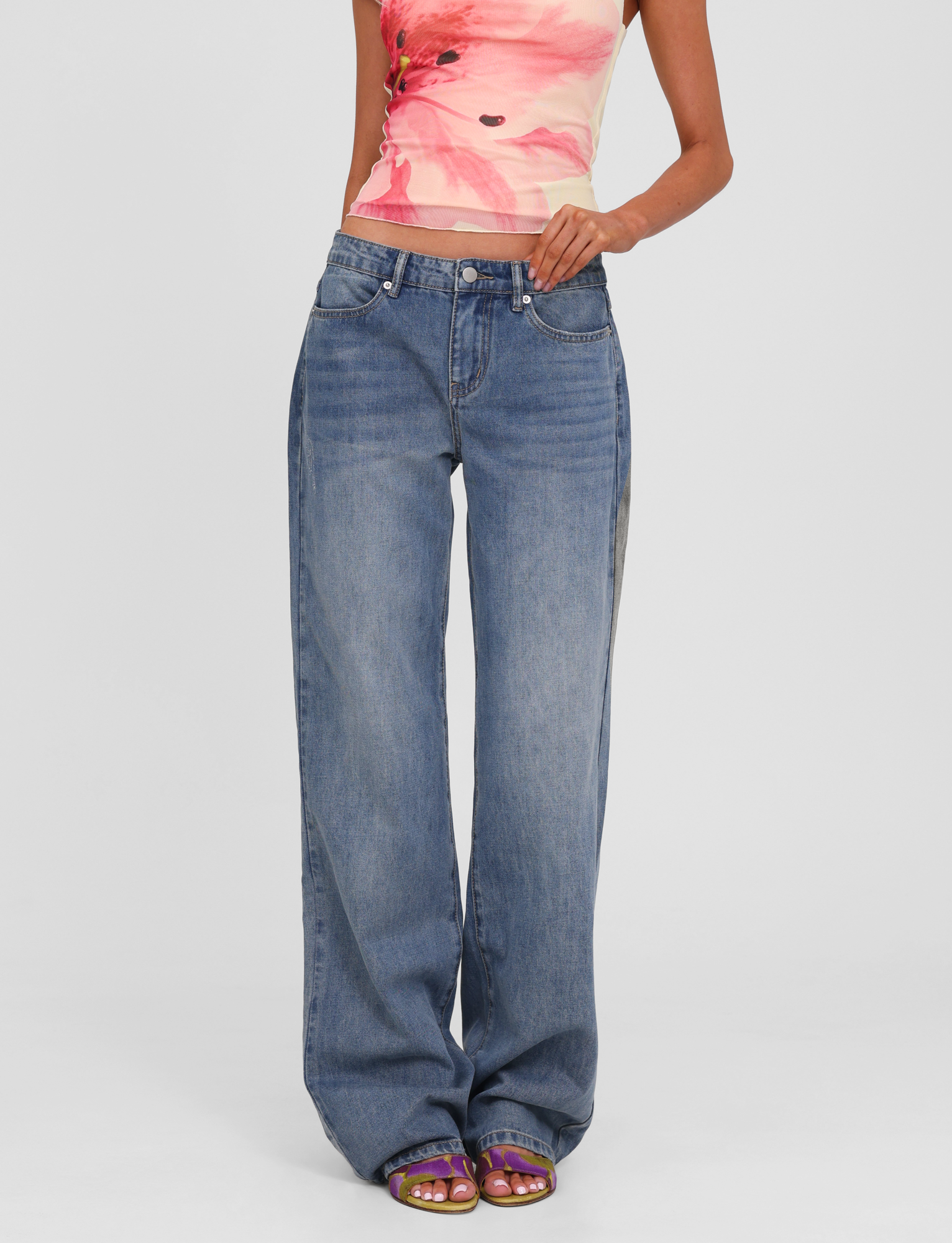 Someone stands against a plain light background wearing TIGERMIST's ESTRELLA JEAN - MID WASH, paired with a colorful sleeveless top with a pink floral print and open-toe purple sandals.